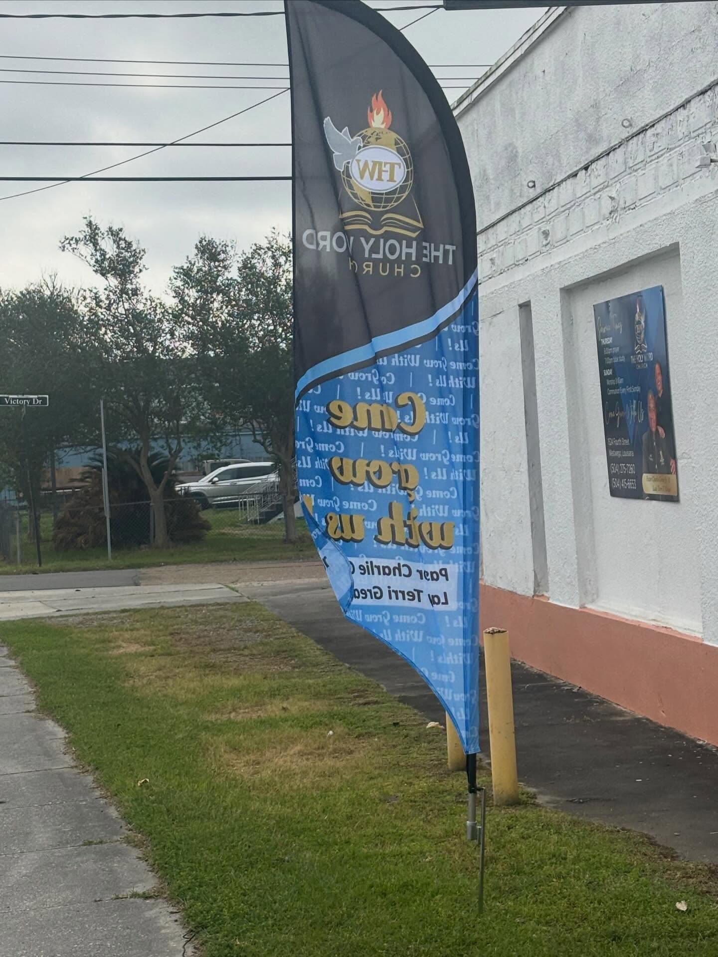 The blue “Come Grow With Us” feather flag outside the white church building at the corner of Victory Drive and 4th Street