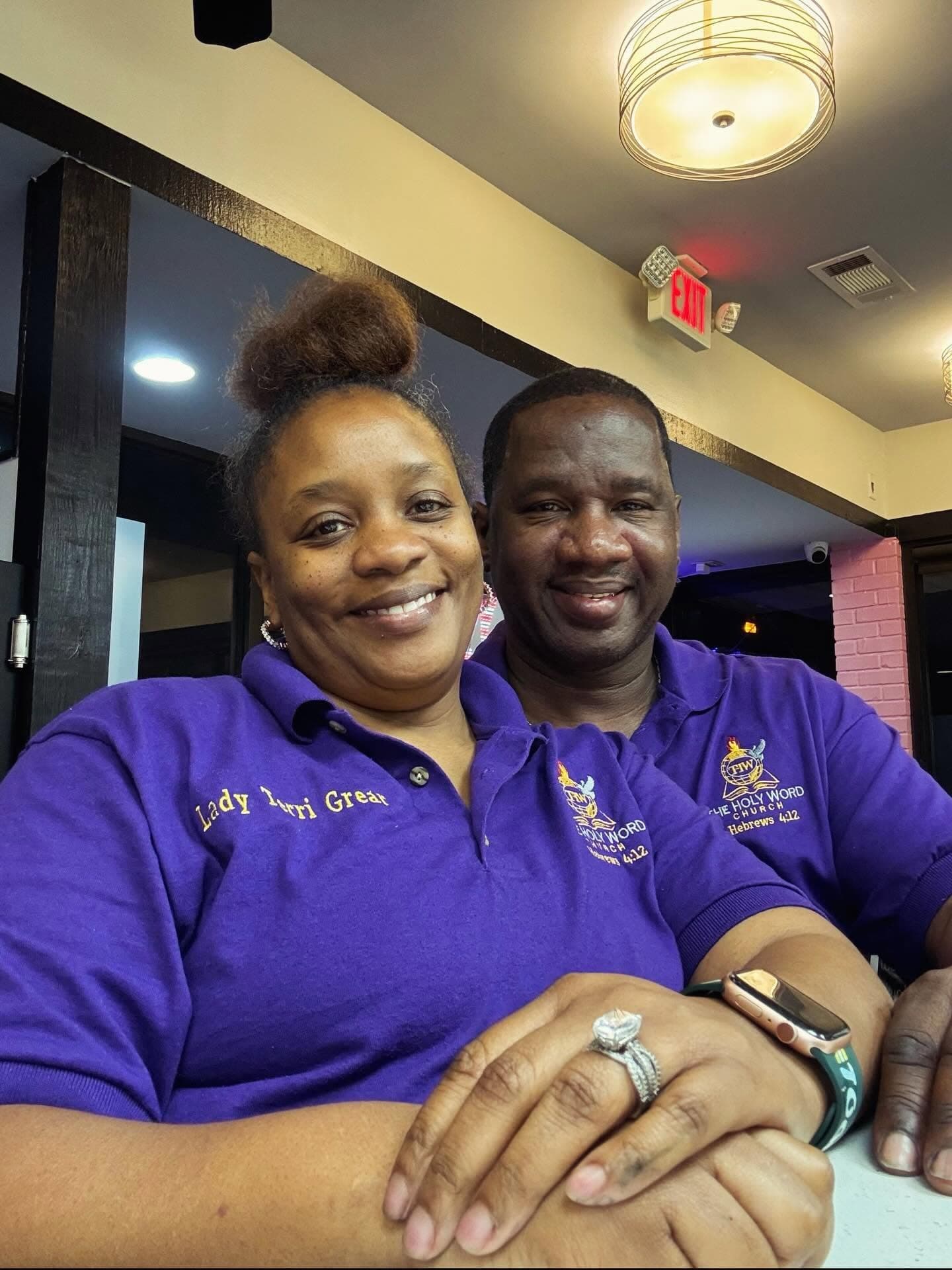 Lady Terri Grear and Pastor Charlie Grear Jr. smiling in purple Holy Word Church polo shirts