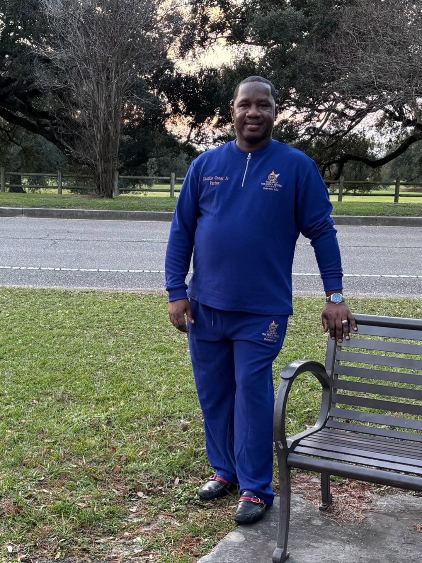 Pastor Charlie Grear Jr. in a royal blue Holy Word Church track suit, standing by a park bench under live oaks